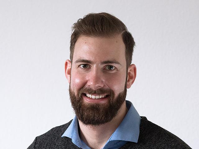 Portrait eines l&auml;chelnden Mannes mit Vollbart, der einen grauen Pullover und ein hellblaues Hemd tr&auml;gt. Helle Wand im Hintergrund.