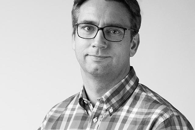 Portr&auml;t eines Mannes mit Brille, der ein kariertes Hemd tr&auml;gt, vor einem neutralen Hintergrund.