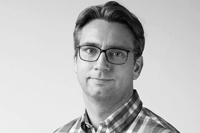 Porträt eines Mannes mit Brille, der ein kariertes Hemd trägt, vor einem neutralen Hintergrund.