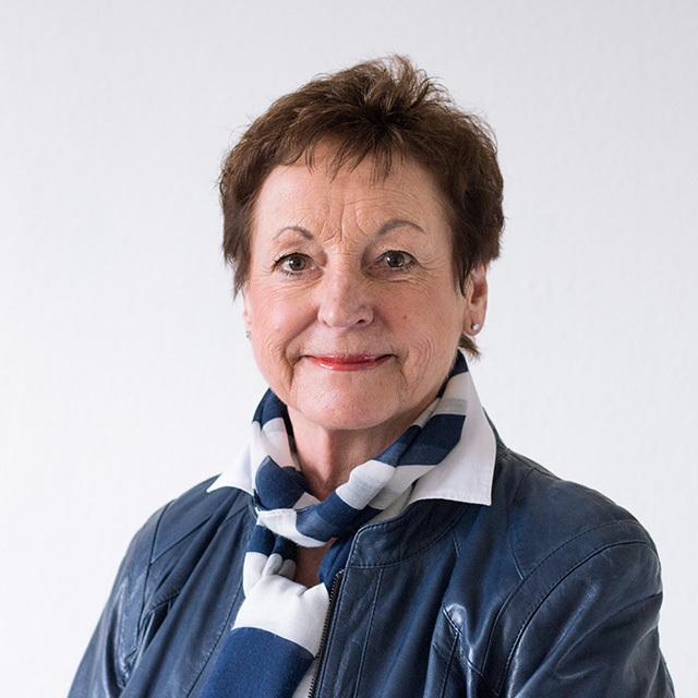 Portr&auml;t einer l&auml;chelnden Frau mit kurzen braunen Haaren, blauer Jacke und gestreiftem Schal vor neutralem Hintergrund.