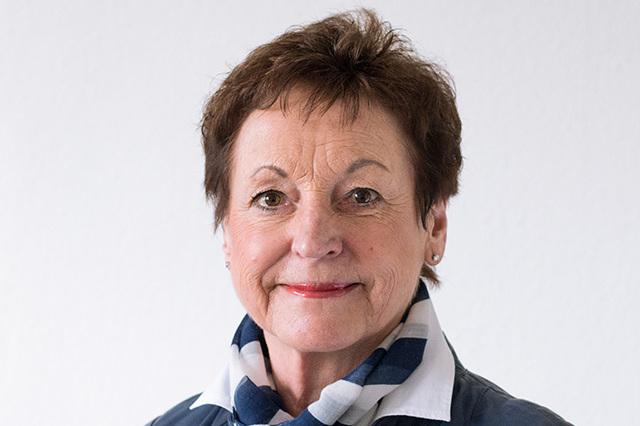 Portr&auml;t einer l&auml;chelnden Frau mit kurzen braunen Haaren, blauer Jacke und gestreiftem Schal vor neutralem Hintergrund.