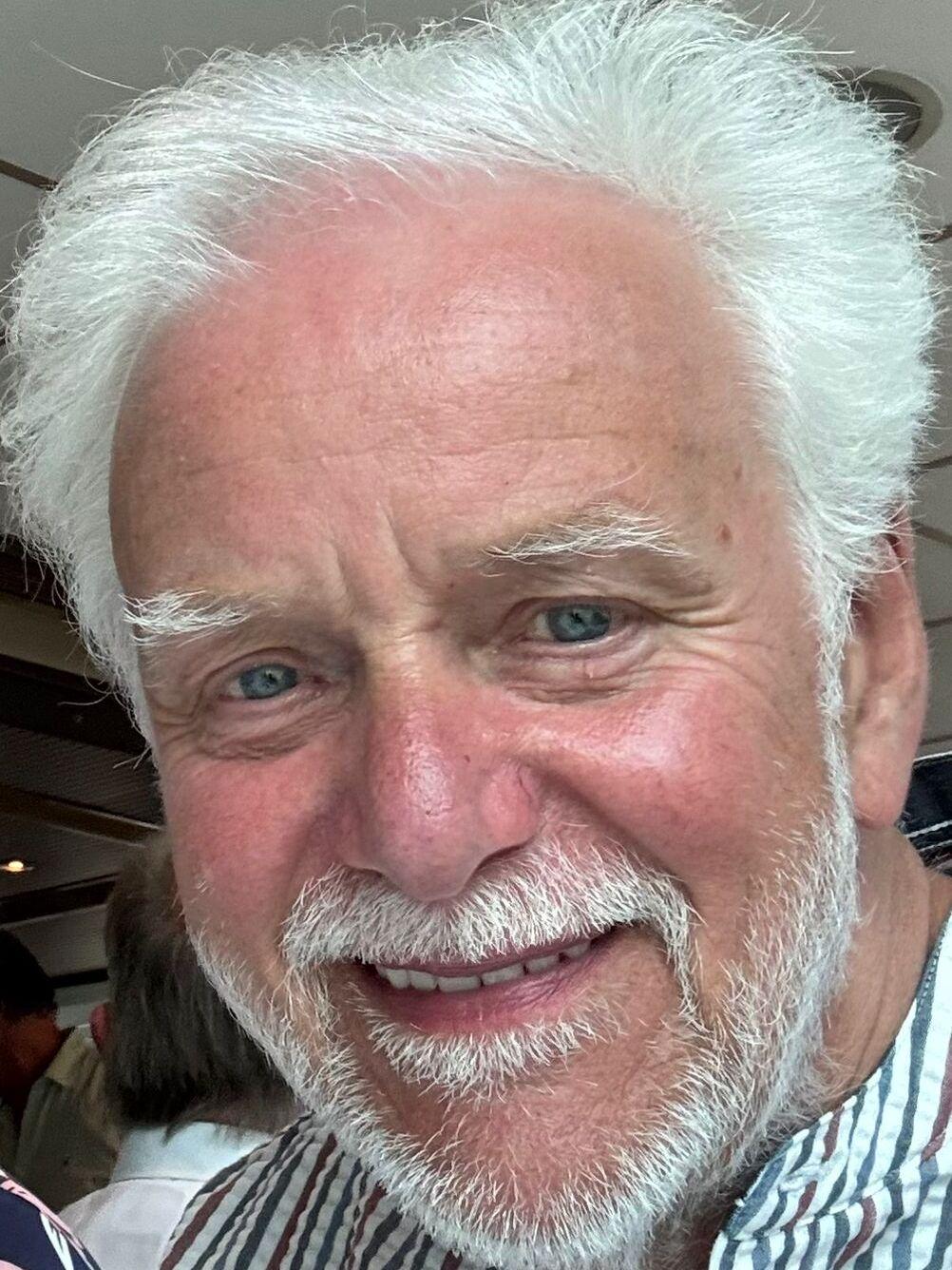 L&auml;chelnder &auml;lterer Mann mit wei&szlig;em Haar und blauem Hemd vor verschwommener Hintergrundszene.