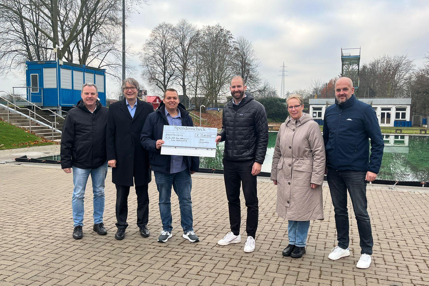 Gruppenzusammenkunft mit Scheck&uuml;bergabe vor einem Schwimmbad, umgeben von B&auml;umen und Geb&auml;uden.