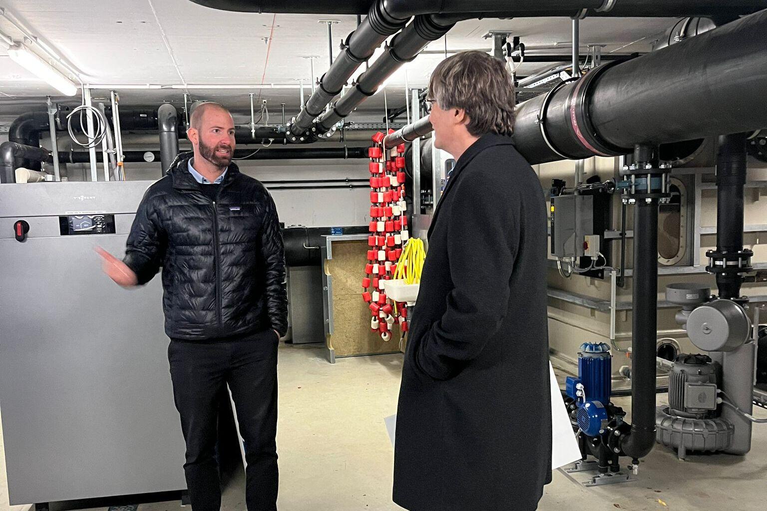 Zwei M&auml;nner im Gespr&auml;ch in einem technischen Raum mit Rohren und Anlagen im Hintergrund.