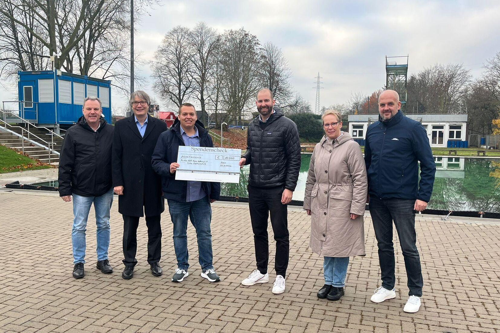 Gruppenzusammenkunft mit Scheck&uuml;bergabe vor einem Schwimmbad, umgeben von B&auml;umen und Geb&auml;uden.