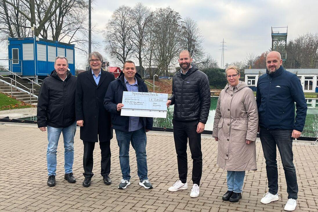 Gruppenzusammenkunft mit Scheck&uuml;bergabe vor einem Schwimmbad, umgeben von B&auml;umen und Geb&auml;uden.