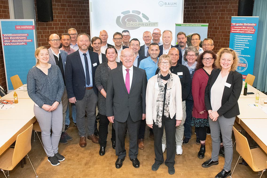 Gruppenfoto von Teilnehmern einer Veranstaltung, l&auml;chelnd vor einer Pr&auml;sentationswand und Tischen in einem Konferenzraum.