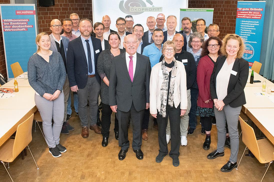 Gruppenfoto von Teilnehmern einer Veranstaltung, l&auml;chelnd vor einer Pr&auml;sentationswand und Tischen in einem Konferenzraum.
