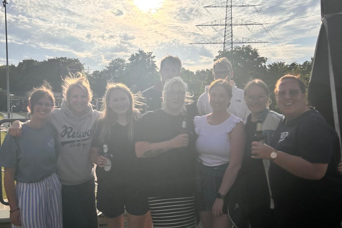 Neun Teilnehmende der Schwimmlehrer-Ausbildung stehen l&auml;chelnd zusammen im Au&szlig;enbereich des Freibads Dellwig. Im Hintergrund sind B&auml;ume, ein Strommast und ein bew&ouml;lkter Abendhimmel mit Sonnenstrahlen zu sehen. Mehrere Personen tragen