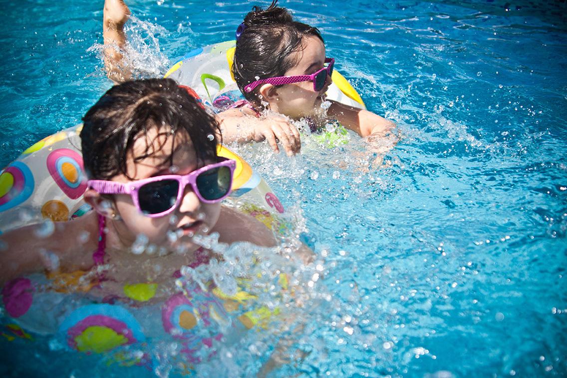 Zwei Mädchen in Schwimmringen mit Sonnenbrillen plantschen im blauen Wasser eines Schwimmbeckens.