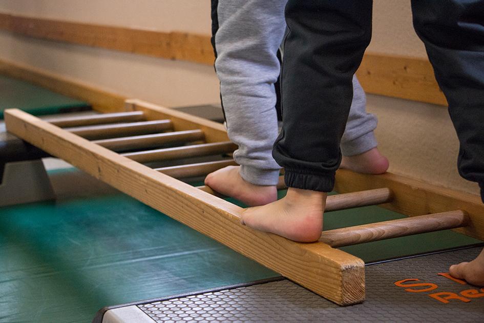 Zwei Barfußfüße balancieren auf einer Holzkletterleiter in einer Turnhalle.