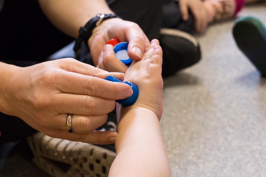 H&auml;nde einer erwachsenen Person platzieren bunte Spielsteine auf dem Handgelenk eines Kleinkindes, w&auml;hrend andere Kinder im Hintergrund sitzen.