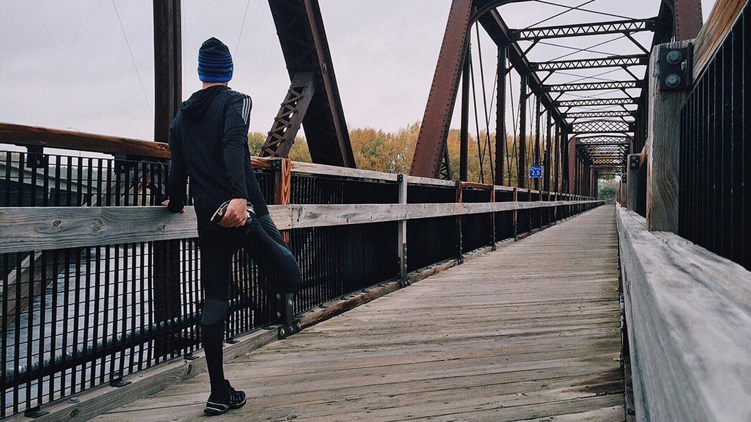 L&auml;ufer in sportlicher Kleidung dehnt sich auf einer Holzbr&uuml;cke mit metallenen Rahmen und herbstlicher Landschaft.