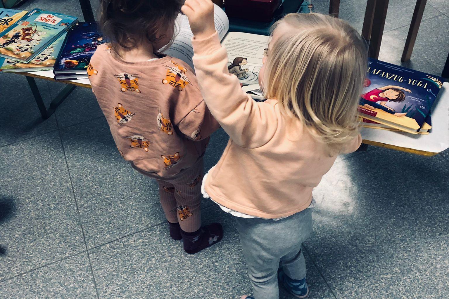 Zwei Kinder stehen vor einem Tisch mit bunten B&uuml;chern und Gegenst&auml;nden, das eine h&auml;lt das andere an der Hand.