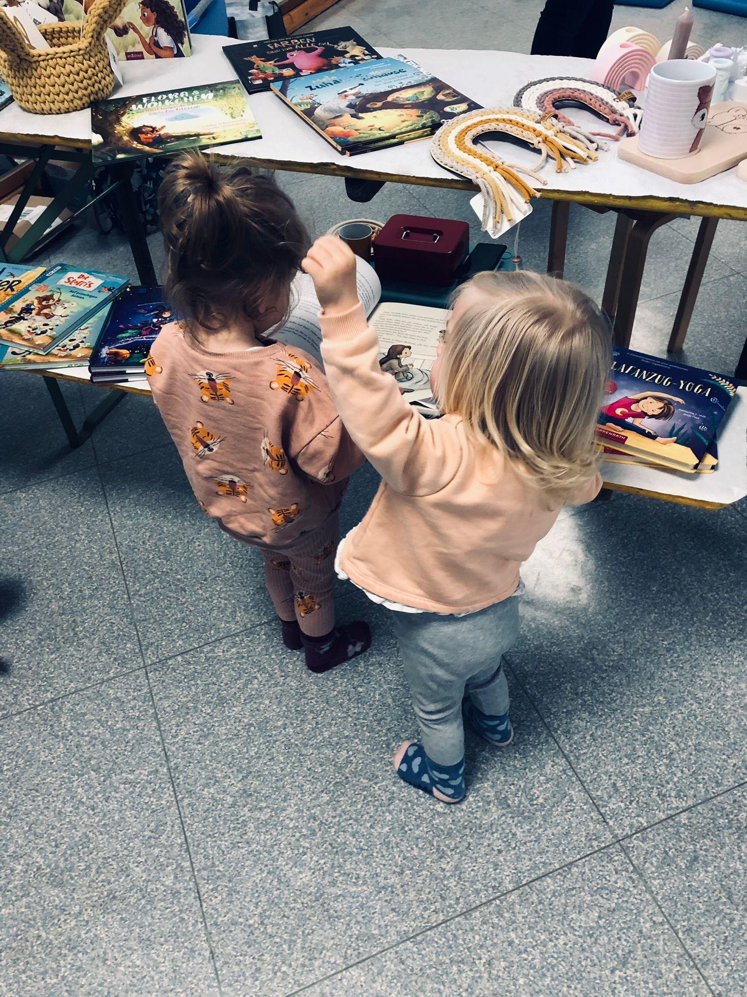 Zwei Kinder stehen vor einem Tisch mit bunten B&uuml;chern und Gegenst&auml;nden, das eine h&auml;lt das andere an der Hand.
