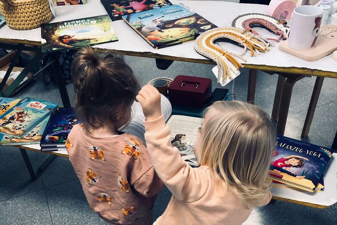 Zwei Kinder stehen vor einem Tisch mit bunten B&uuml;chern und Gegenst&auml;nden, das eine h&auml;lt das andere an der Hand.