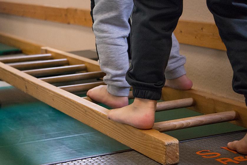 Zwei barfüßige Kinder stehen auf einer Holzleiter, die auf einer Gymnastikmatte platziert ist.