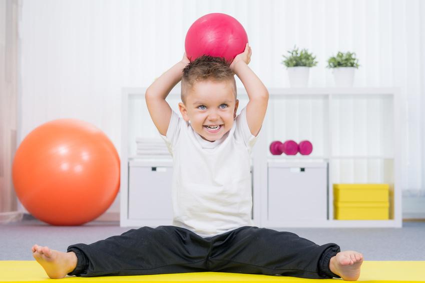 Junge sitzt mit gespreizten Beinen, hebt einen roten Ball und lächelt. Gymnastikbälle und Regale im Hintergrund.