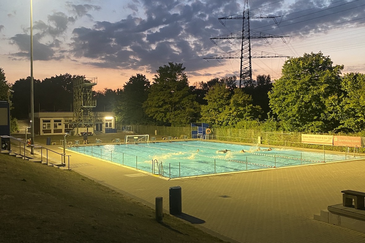 Beleuchtetes Schwimmbecken im Freien bei Sonnenuntergang, umgeben von B&auml;umen und Wolken.