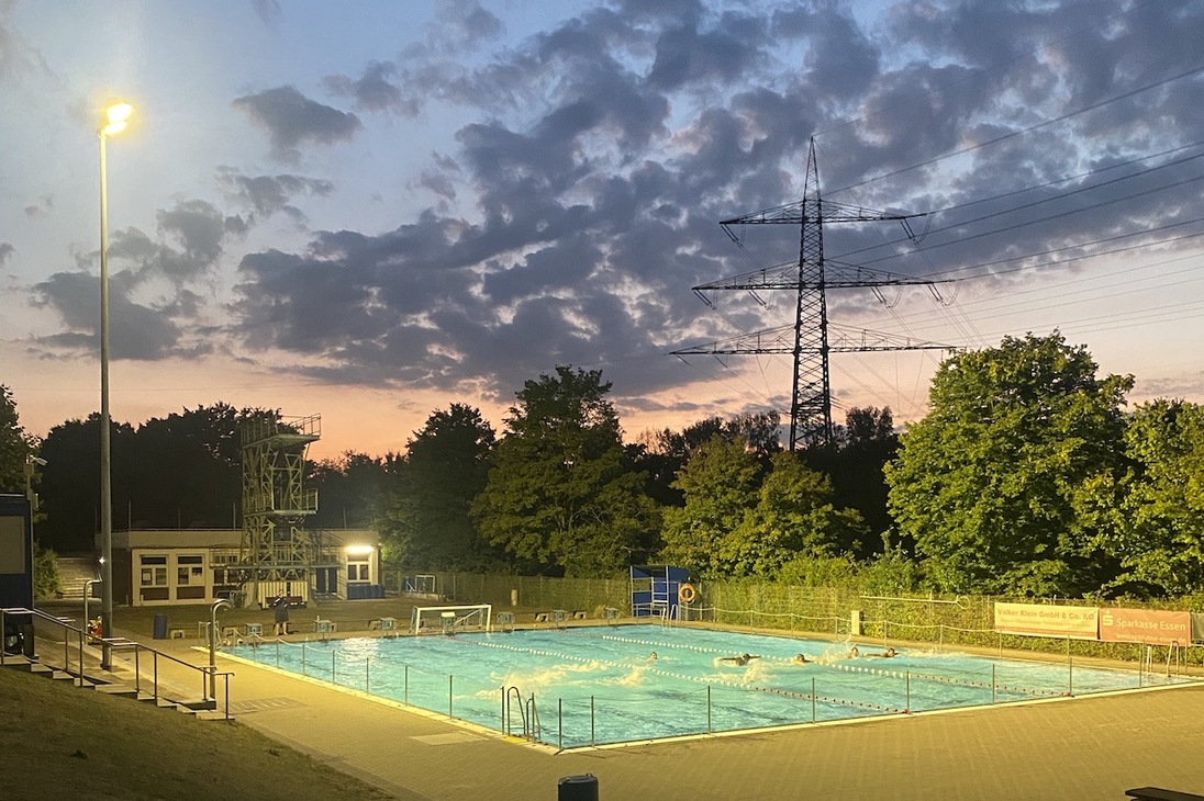 Beleuchtetes Schwimmbecken im Freien bei Sonnenuntergang, umgeben von B&auml;umen und Wolken.