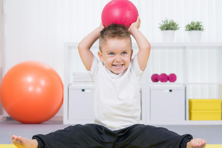 Junge sitzt mit gespreizten Beinen, hebt einen roten Ball und l&auml;chelt. Gymnastikb&auml;lle und Regale im Hintergrund.
