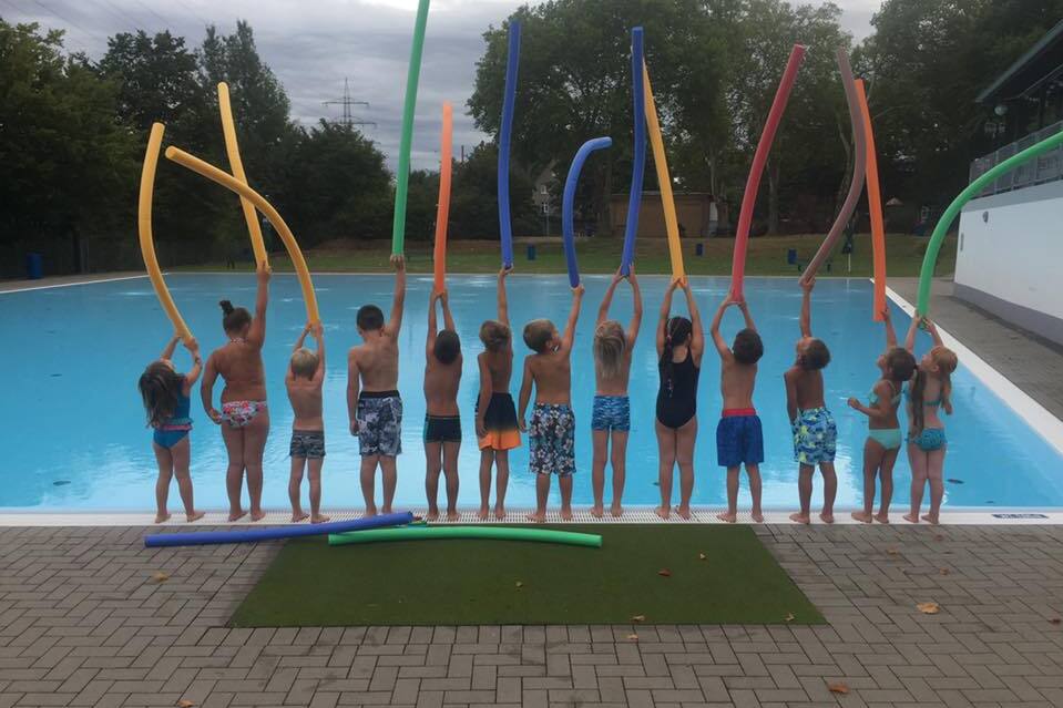 Gruppe von Kindern steht am Pool und h&auml;lt farbige Schwimmnudeln in die H&ouml;he.