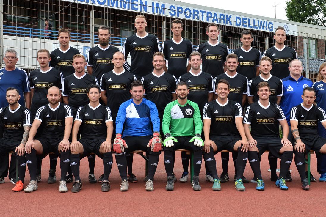 Gruppenfoto einer Fu&szlig;ballmannschaft in schwarz-wei&szlig;er Sportkleidung im Sportpark Dellwig vor einem Begr&uuml;&szlig;ungsschild.