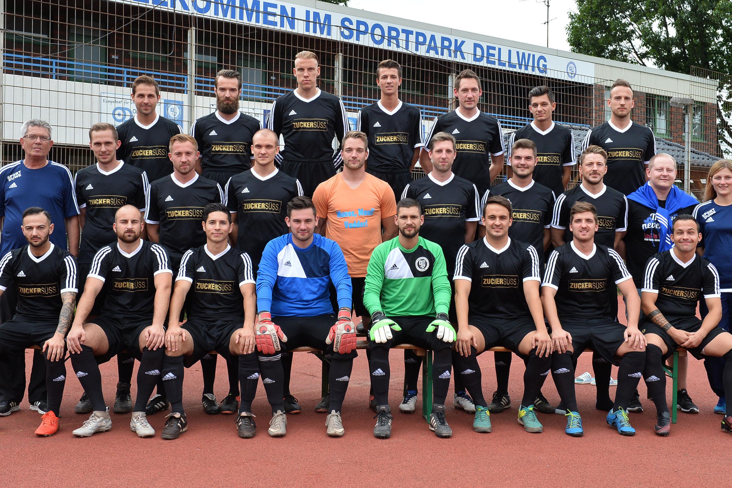 Mannschaftsfoto einer Fu&szlig;ballmannschaft in schwarzen Trikots, stehend und sitzend, vor einem Sportpark.