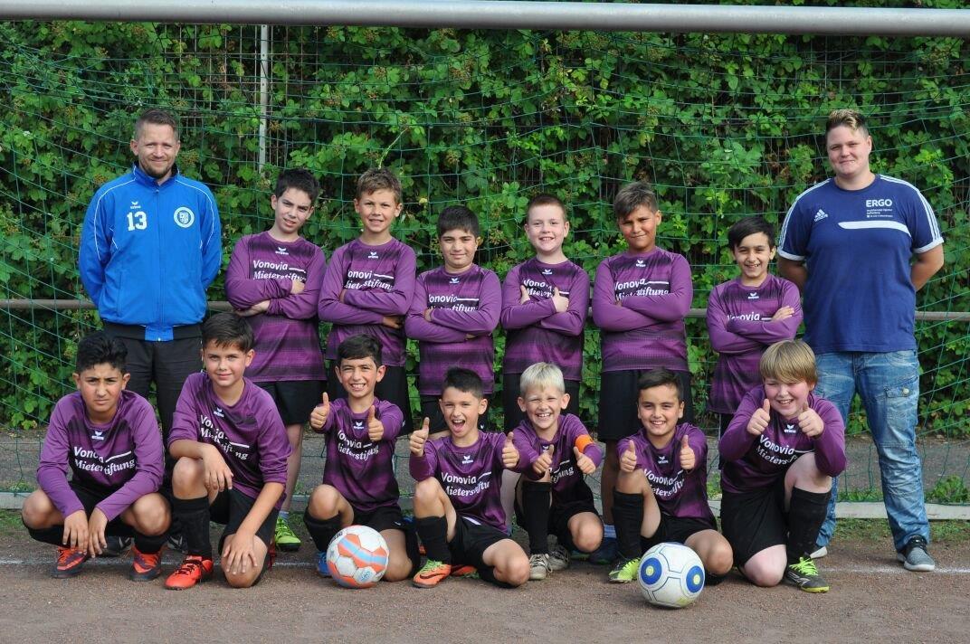 Fußballmannschaft von zwölf Kindern in lila Trikots, posierend mit drei Bällen und zwei Trainern auf einem Sportplatz.