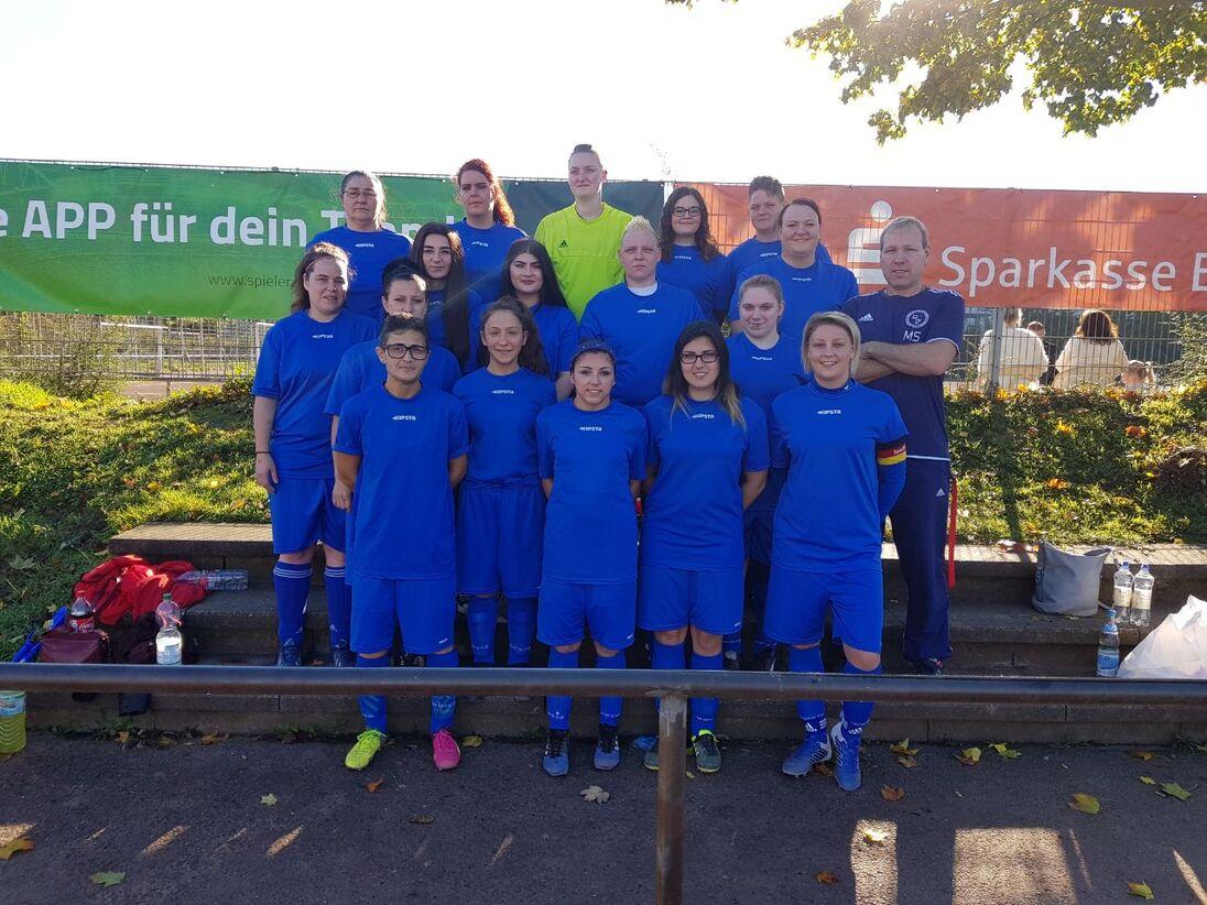 Frauenfußballmannschaft in blauen Trikots steht vor einer grünen Werbetafel und lächelt in die Kamera.