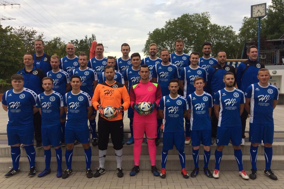 Gruppenfoto einer Fu&szlig;ballmannschaft in blauen Trikots, einschlie&szlig;lich Torh&uuml;tern, auf einem Platz mit B&auml;umen im Hintergrund.