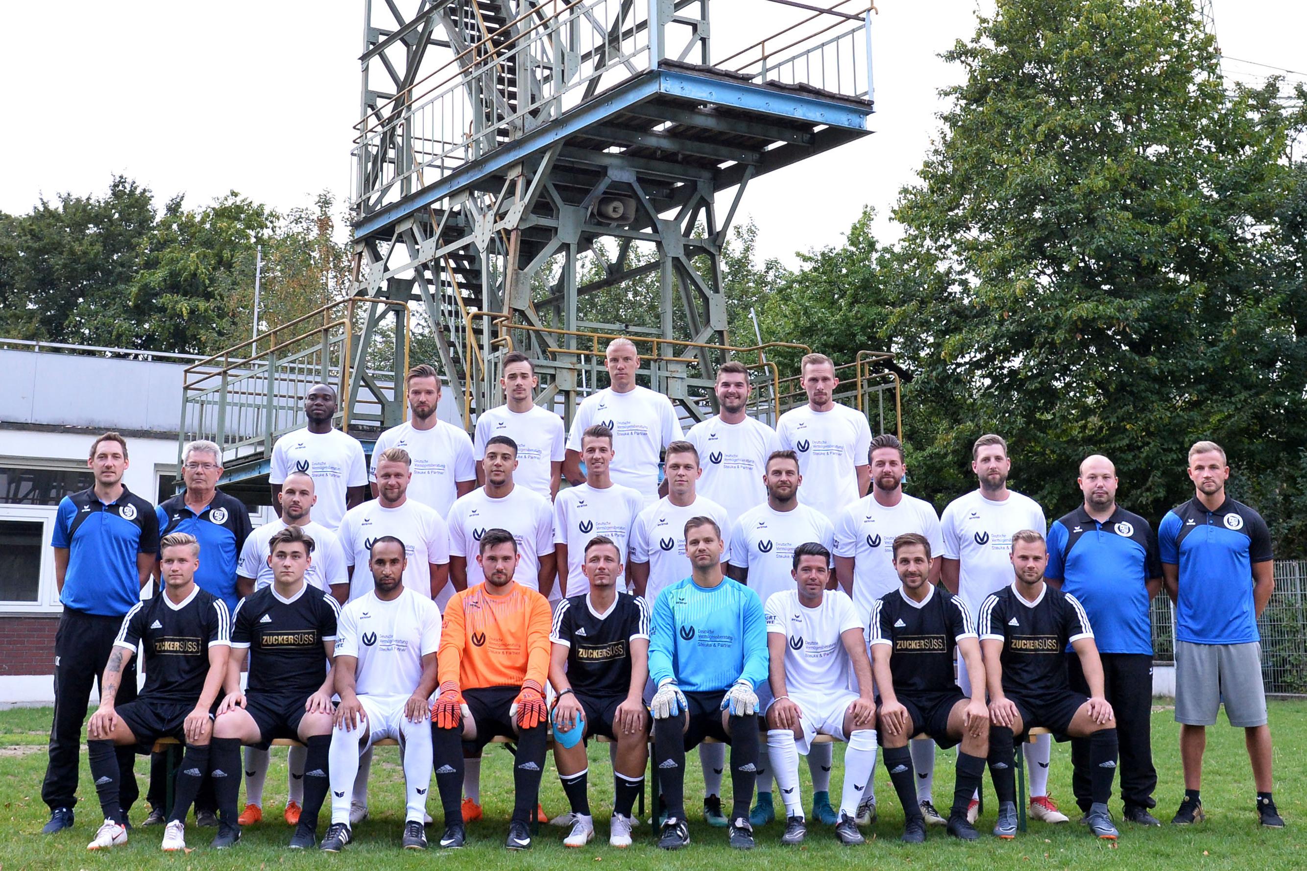 Mannschaftsfoto einer Fu&szlig;ballmannschaft mit 25 Spielern in Trikots sowie Trainer unter einem F&ouml;rderturm im Hintergrund.