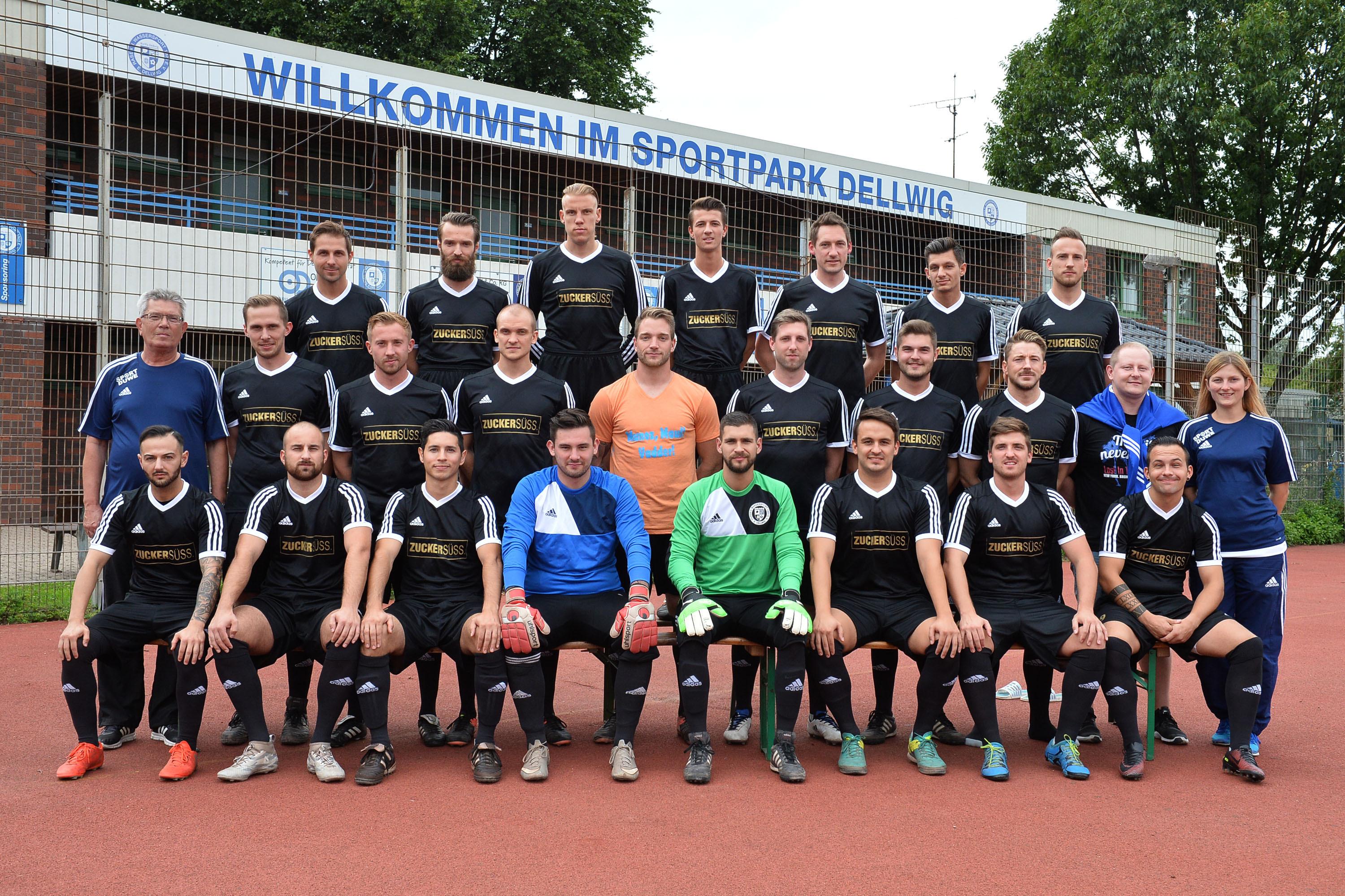 Mannschaftsfoto einer Fu&szlig;ballmannschaft in schwarzen Trikots, stehend und sitzend, vor einem Sportpark.