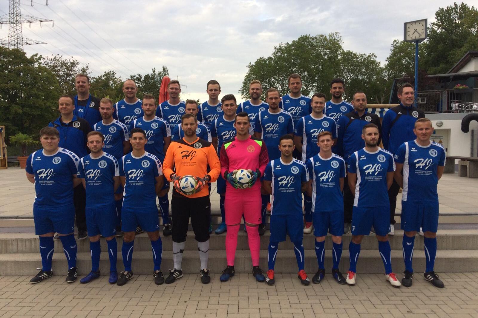 Gruppenfoto einer Fu&szlig;ballmannschaft in blauen Trikots, einschlie&szlig;lich Torh&uuml;tern, auf einem Platz mit B&auml;umen im Hintergrund.