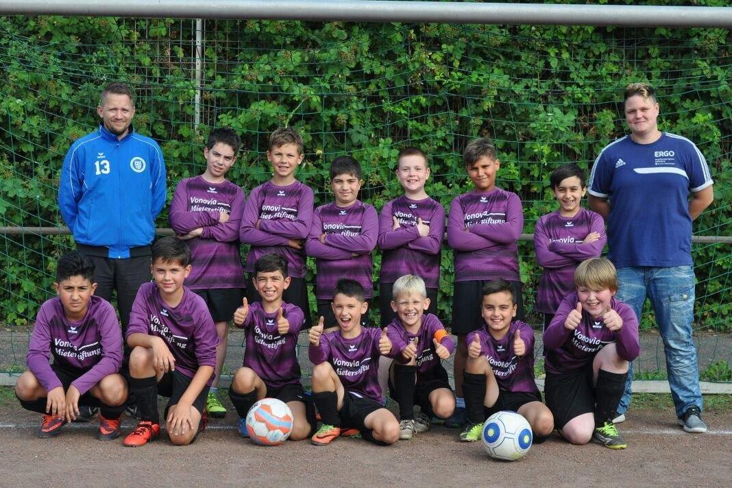 Fu&szlig;ballmannschaft von zw&ouml;lf Kindern in lila Trikots, posierend mit drei B&auml;llen und zwei Trainern auf einem Sportplatz.