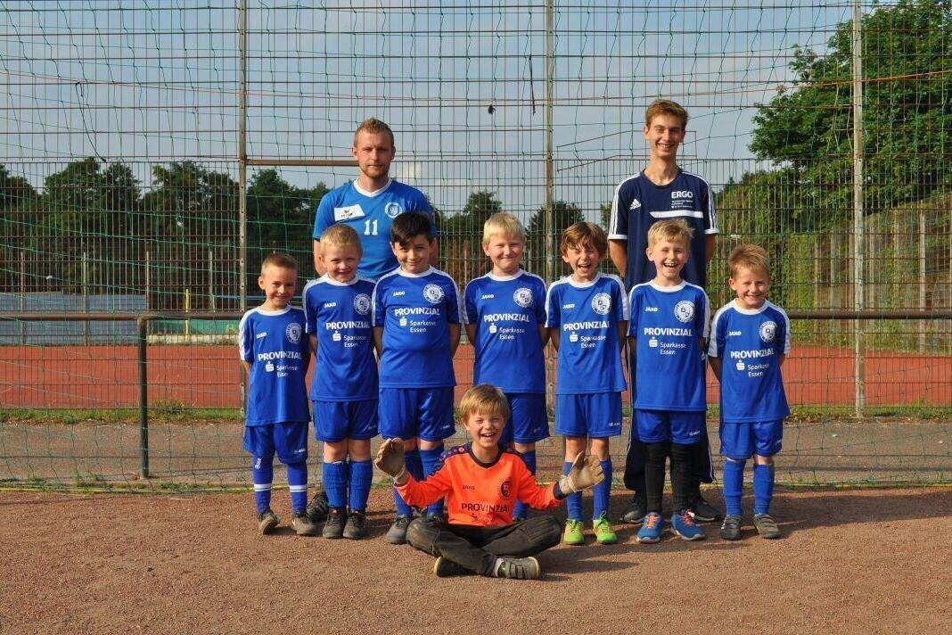 Kinderfu&szlig;ballmannschaft mit Trainern in blauen Trikots, stehend und sitzend im Sportpark hinter einem Tornetz.