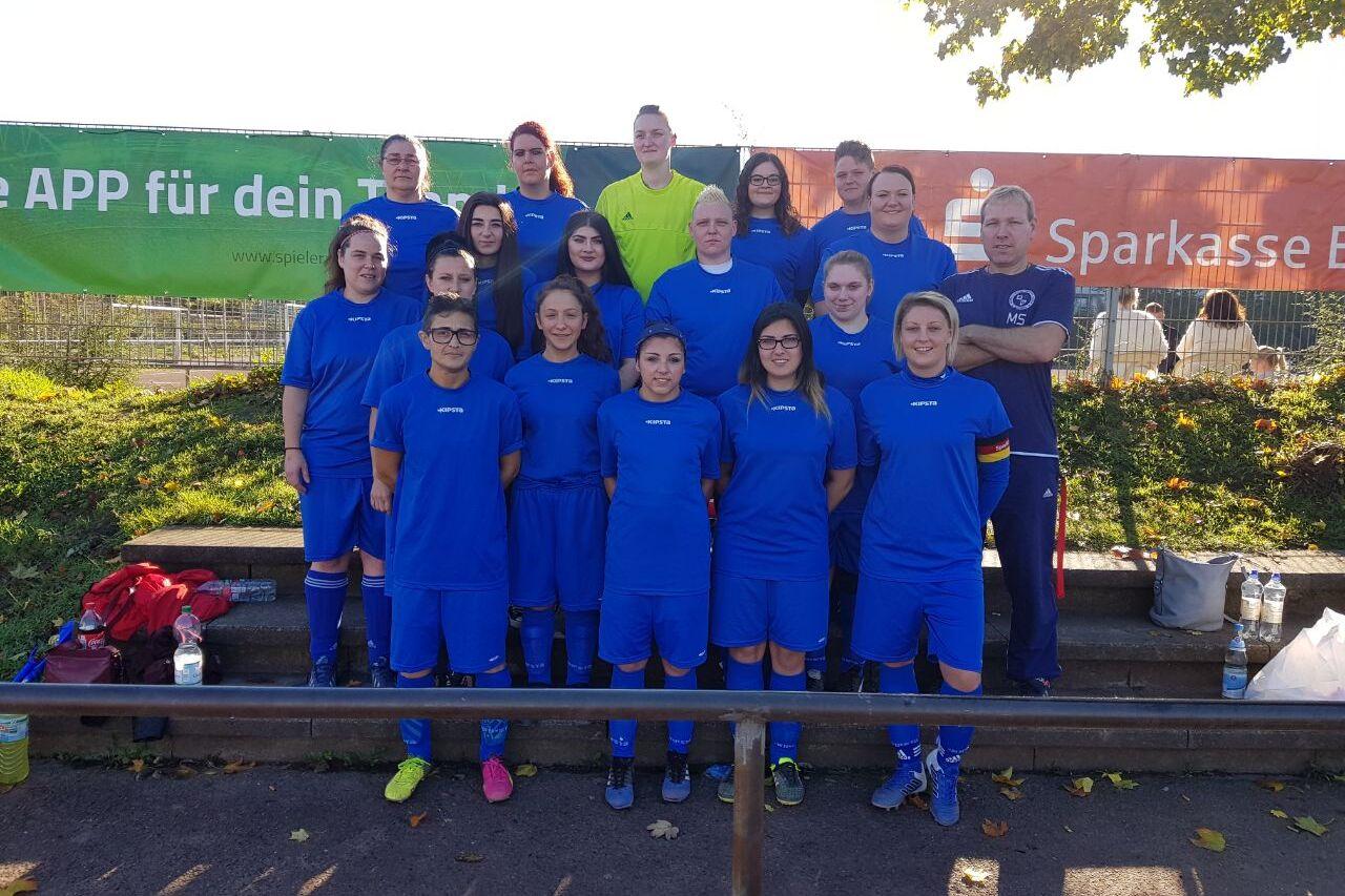 Frauenfu&szlig;ballmannschaft in blauen Trikots steht vor einer gr&uuml;nen Werbetafel und l&auml;chelt in die Kamera.