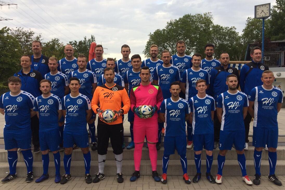 Gruppenfoto einer Fu&szlig;ballmannschaft in blauen Trikots, einschlie&szlig;lich Torh&uuml;tern, auf einem Platz mit B&auml;umen im Hintergrund.