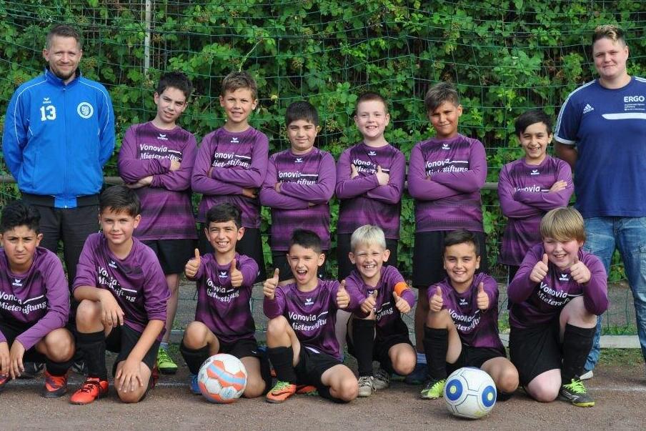 Fußballmannschaft von zwölf Kindern in lila Trikots, posierend mit drei Bällen und zwei Trainern auf einem Sportplatz.