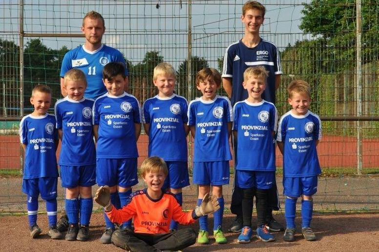 Kinderfu&szlig;ballmannschaft mit Trainern in blauen Trikots, stehend und sitzend im Sportpark hinter einem Tornetz.