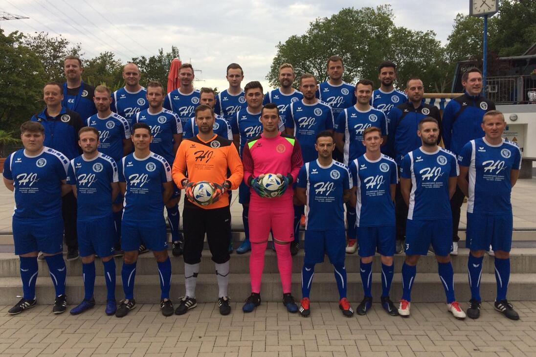 Gruppenfoto einer Fu&szlig;ballmannschaft in blauen Trikots, einschlie&szlig;lich Torh&uuml;tern, auf einem Platz mit B&auml;umen im Hintergrund.