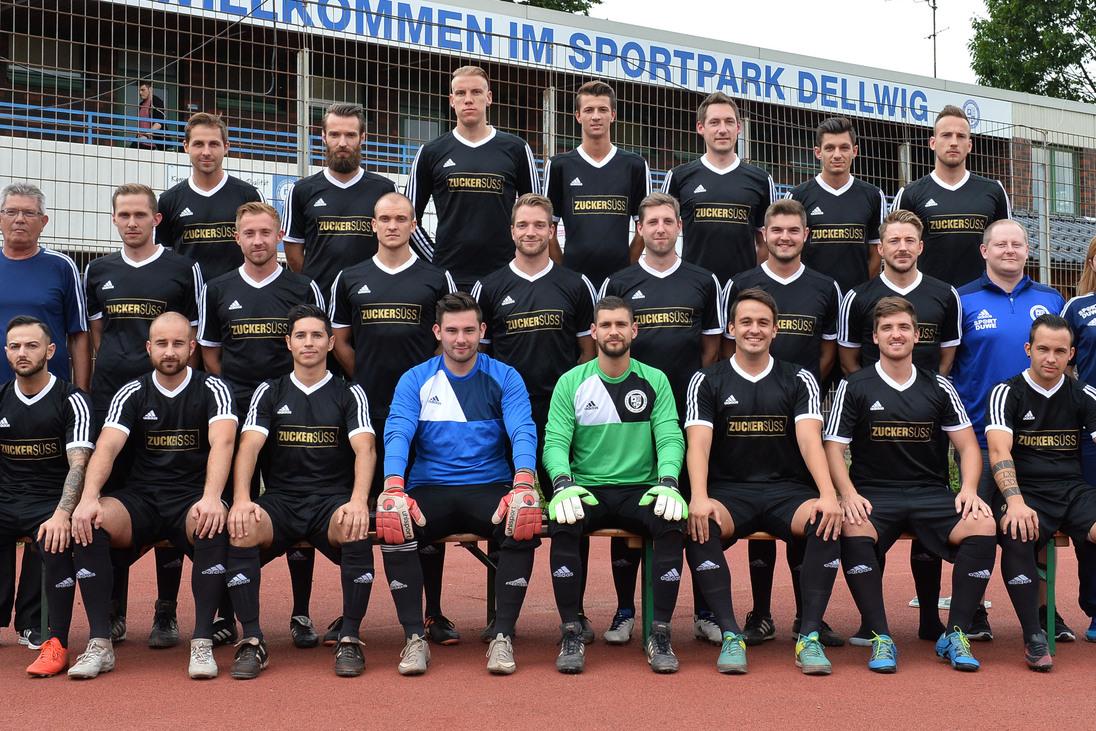 Gruppenfoto einer Fu&szlig;ballmannschaft in schwarz-wei&szlig;er Sportkleidung im Sportpark Dellwig vor einem Begr&uuml;&szlig;ungsschild.