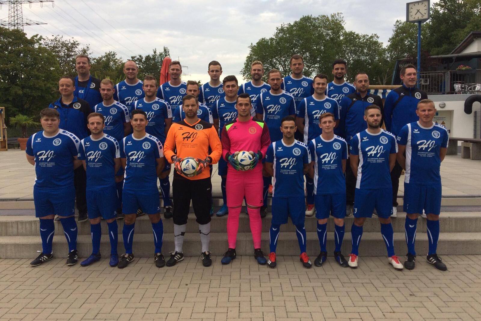 Gruppenfoto einer Fußballmannschaft in blauen Trikots, einschließlich Torhütern, auf einem Platz mit Bäumen im Hintergrund.