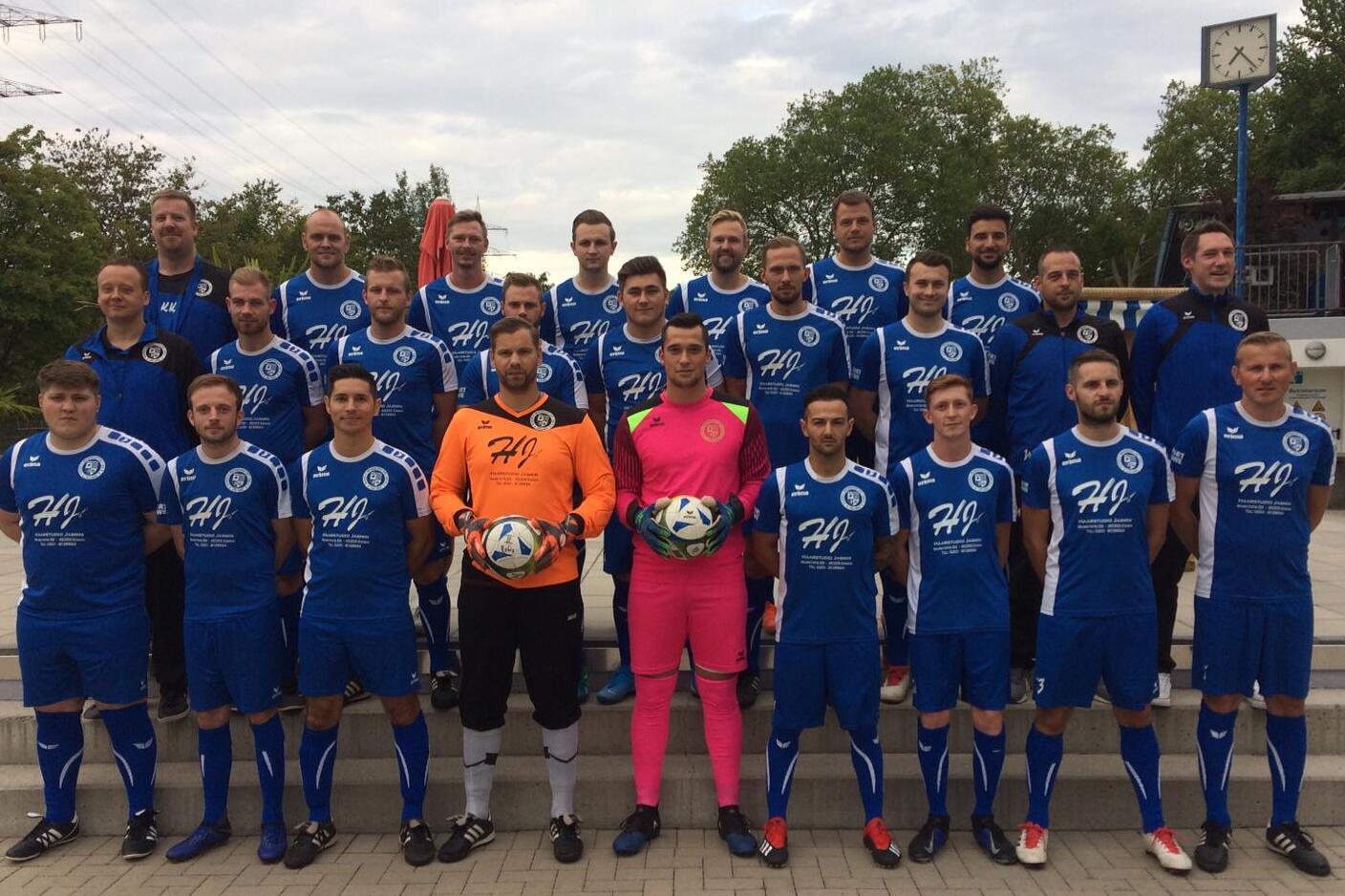Gruppenfoto einer Fußballmannschaft in blauen Trikots, einschließlich Torhütern, auf einem Platz mit Bäumen im Hintergrund.