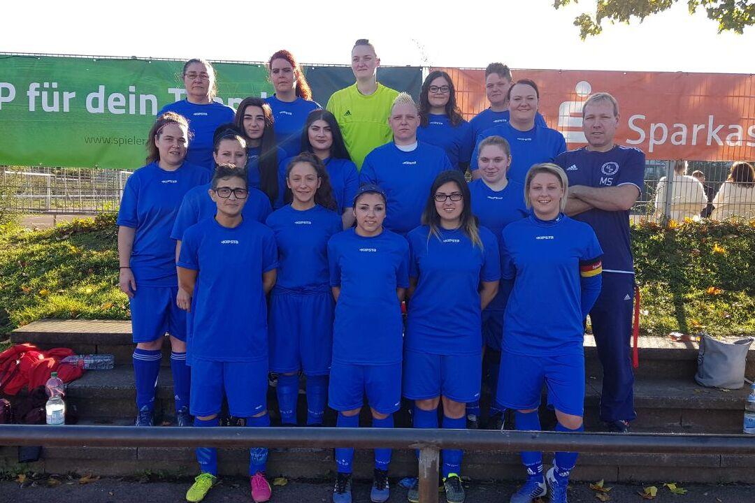 Frauenfußballmannschaft in blauen Trikots steht vor einer grünen Werbetafel und lächelt in die Kamera.