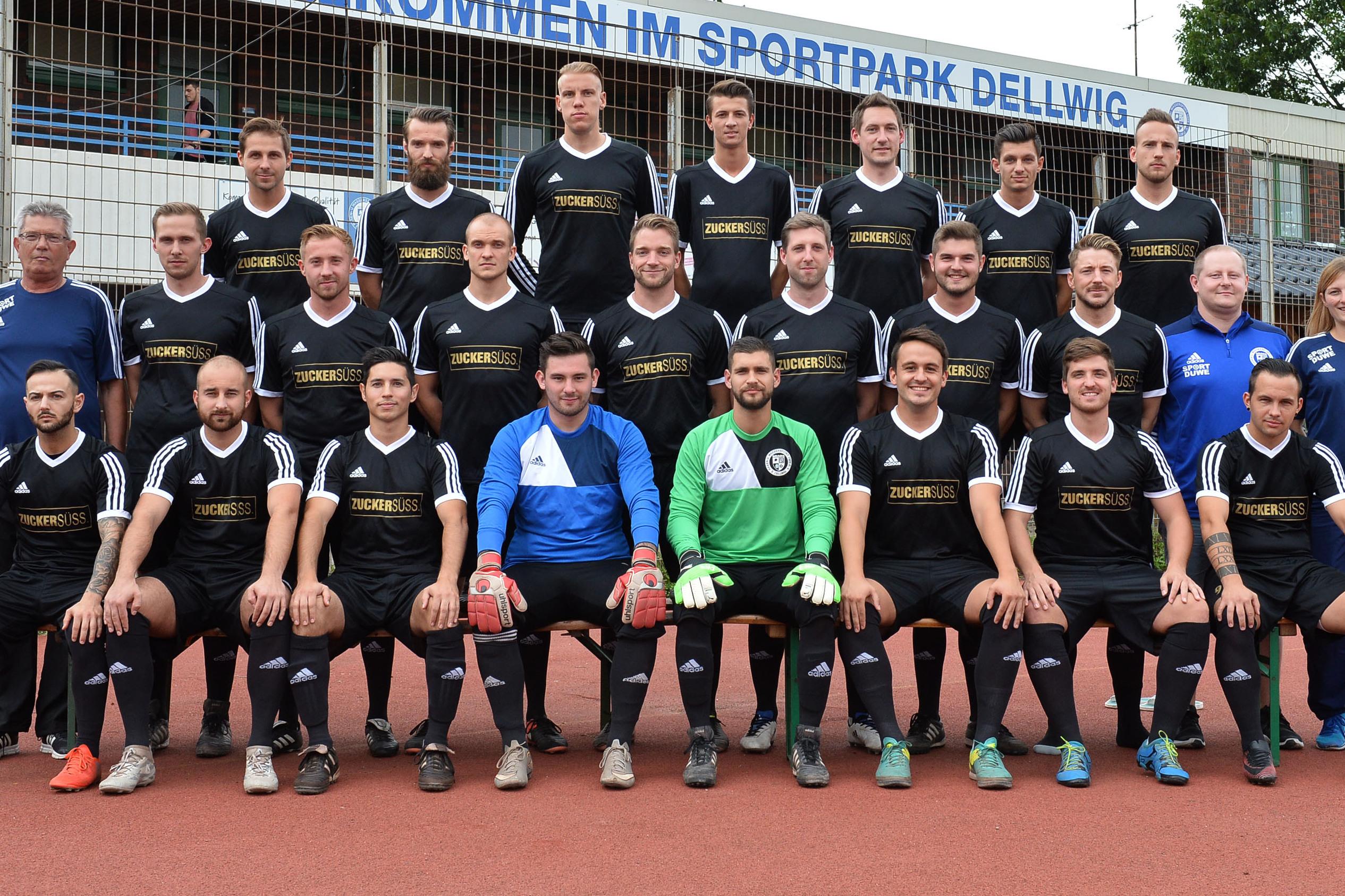 Gruppenfoto einer Fu&szlig;ballmannschaft in schwarz-wei&szlig;er Sportkleidung im Sportpark Dellwig vor einem Begr&uuml;&szlig;ungsschild.
