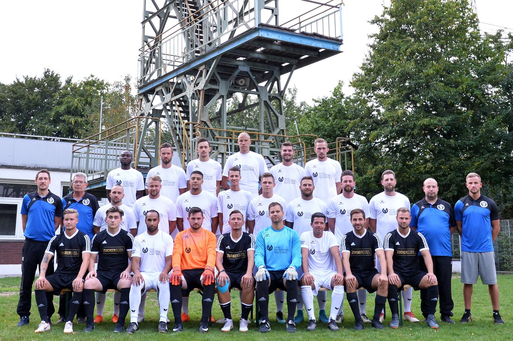 Mannschaftsfoto einer Fu&szlig;ballmannschaft mit 25 Spielern in Trikots sowie Trainer unter einem F&ouml;rderturm im Hintergrund.
