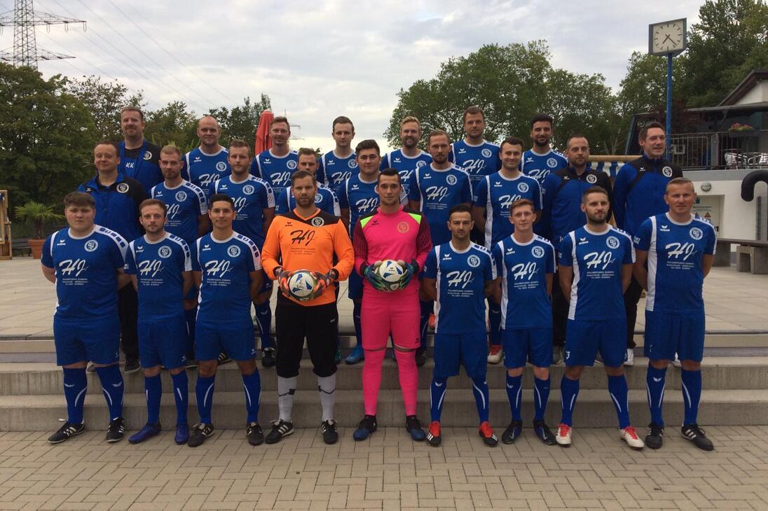 Gruppenfoto einer Fu&szlig;ballmannschaft in blauen Trikots, einschlie&szlig;lich Torh&uuml;tern, auf einem Platz mit B&auml;umen im Hintergrund.