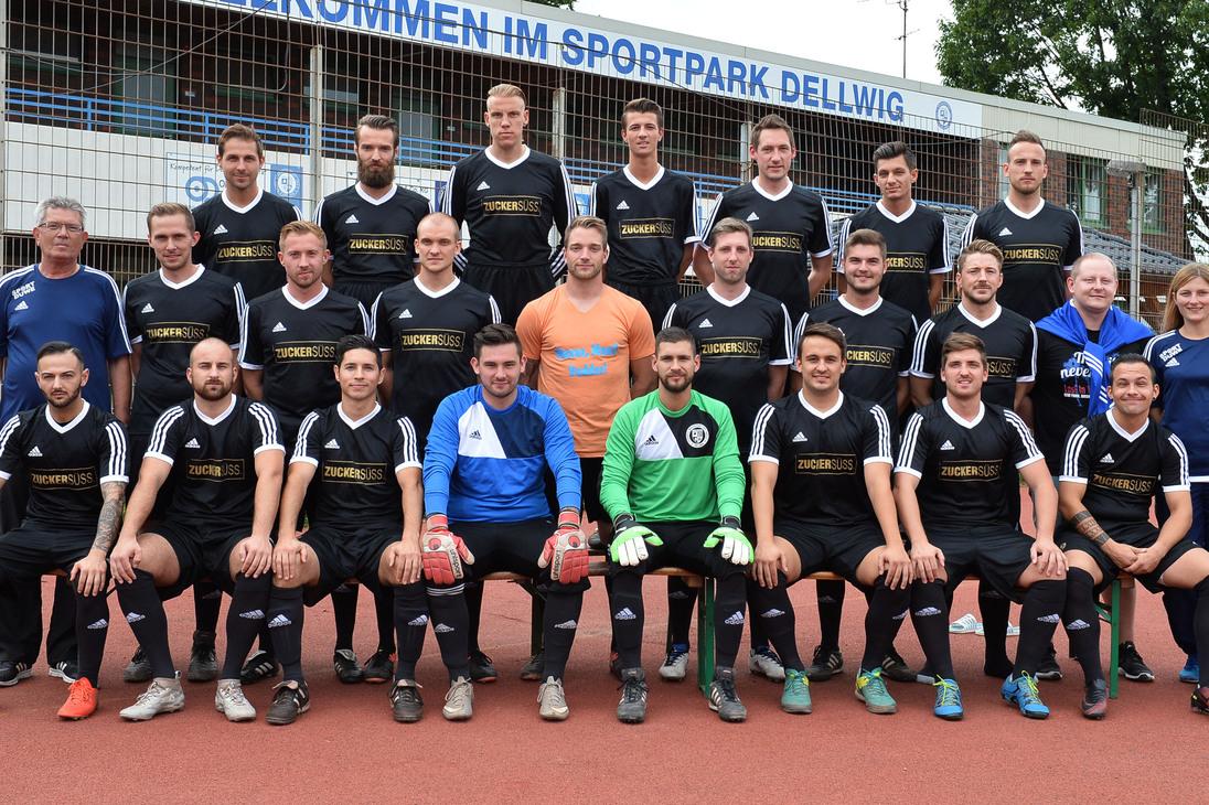 Mannschaftsfoto einer Fu&szlig;ballmannschaft in schwarzen Trikots, stehend und sitzend, vor einem Sportpark.