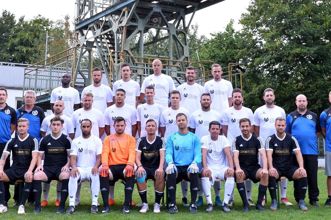 Mannschaftsfoto einer Fu&szlig;ballmannschaft mit 25 Spielern in Trikots sowie Trainer unter einem F&ouml;rderturm im Hintergrund.